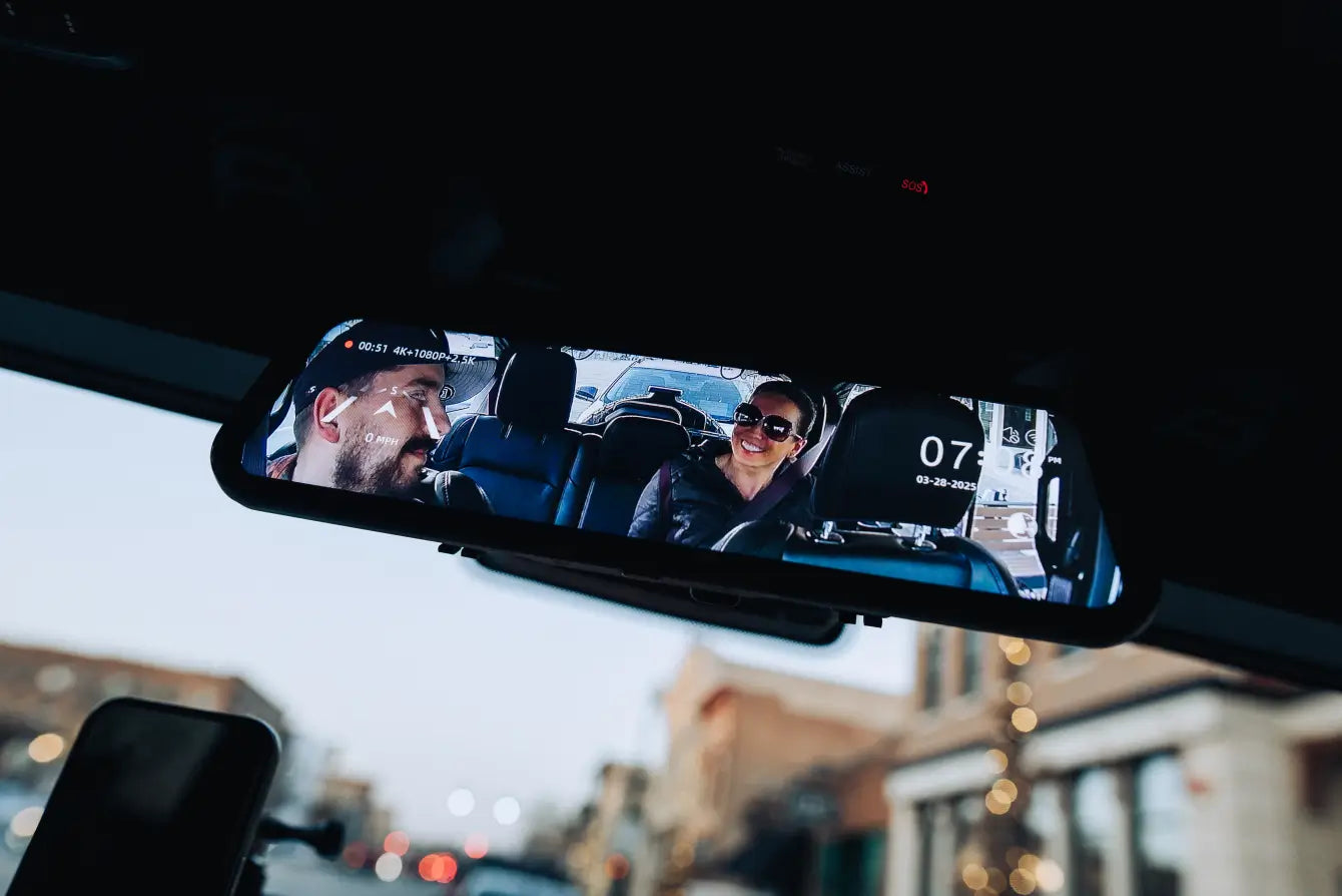 A rearview mirror car radar detector showing a live traffic camera feed in a vehicle interior, with a driver and passenger visible on the screen.