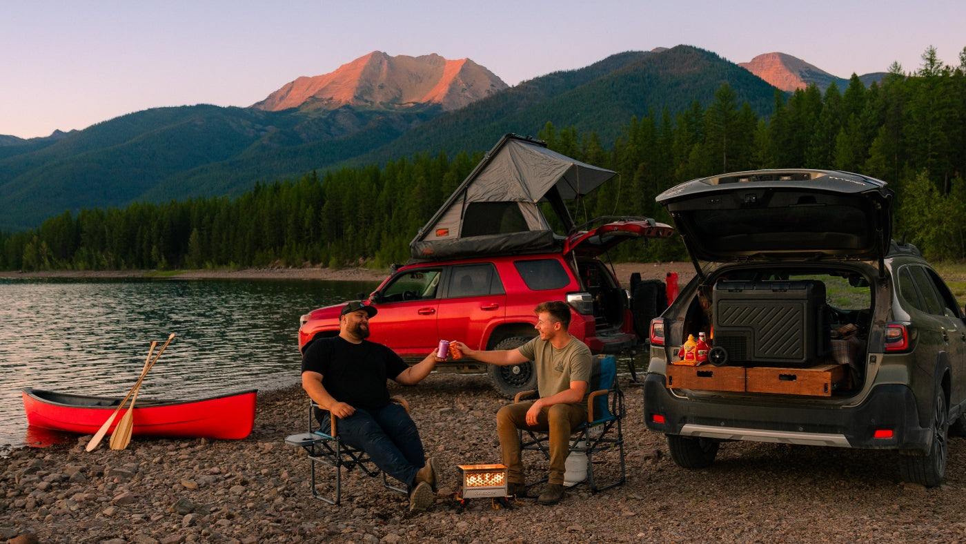 Two men cheering with drinks while car camping by a scenic lakefront with mountains.