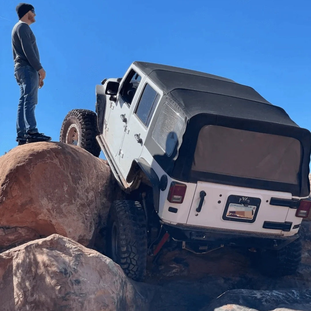 White Jeep Wrangler rock crawling on large desert boulders with a spotter guiding the vehicle.